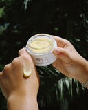 Open jar of Apér Labs The Brightening Butter held in two hands, with a swatch of the balm on the back of one hand